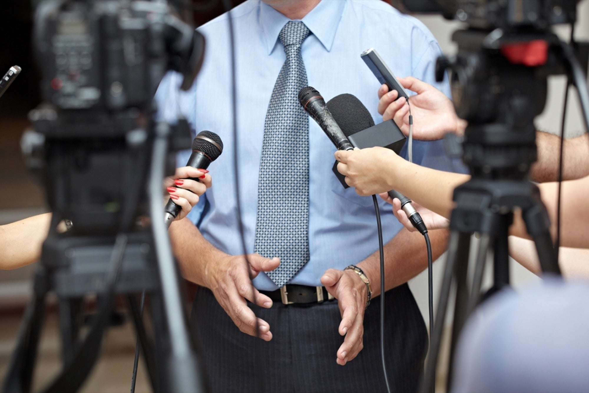 Man wearing blue shirt and tie being interviewed with mics and cameras in front of him