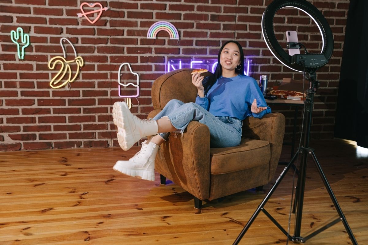 A girl seating in a sofa eating pizza with ring light and phone in front of her