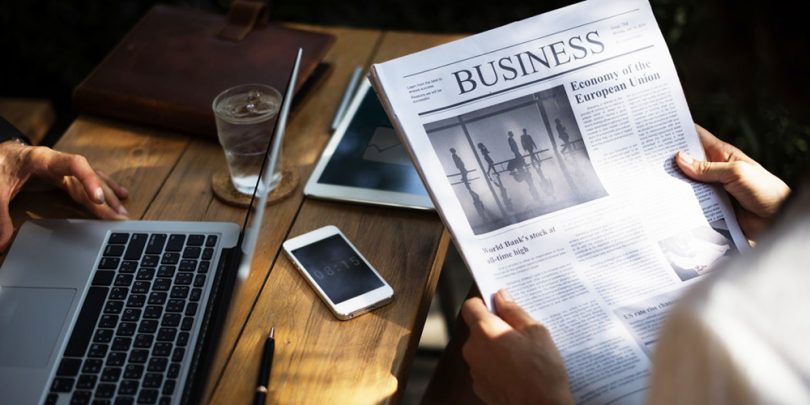 A newspaper with word 'business' as heading, a laptop, phone, tablet, and glass of water on desk