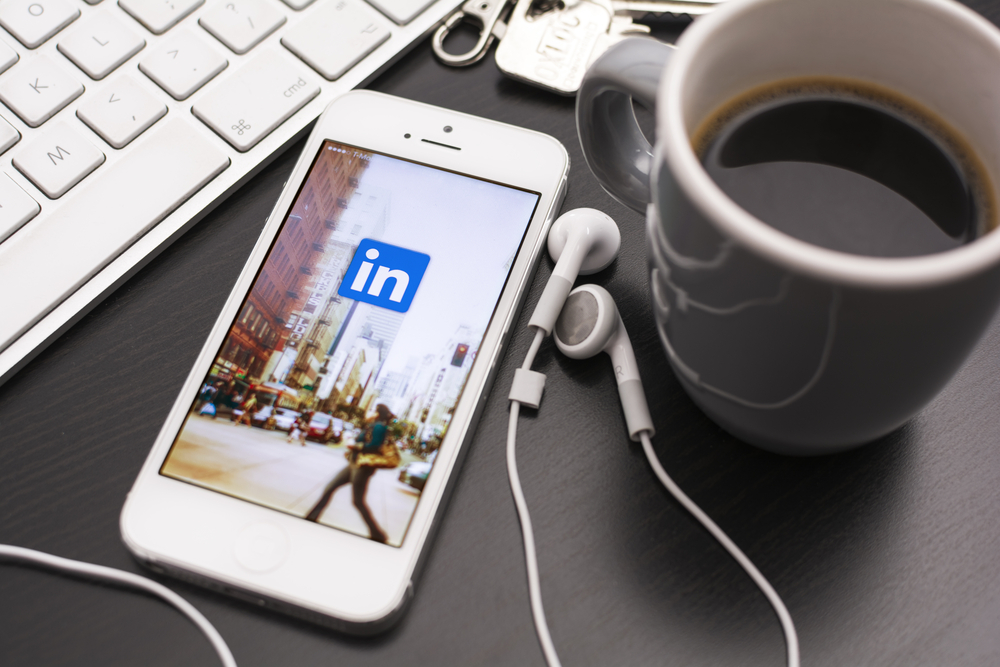 LinkedIn logo in phone's screen with mug, hearphones, and keyboard beside it
