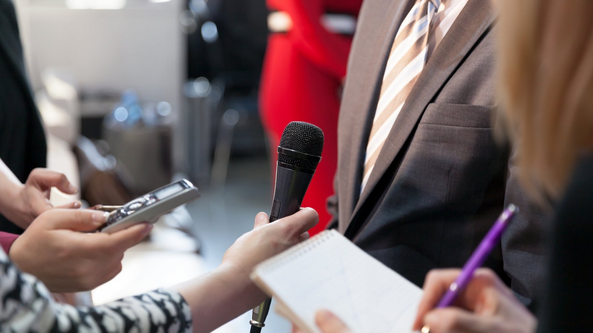 Reporters holding microphones, voice recorder, pen and notebbok while interviewing a man in a suit