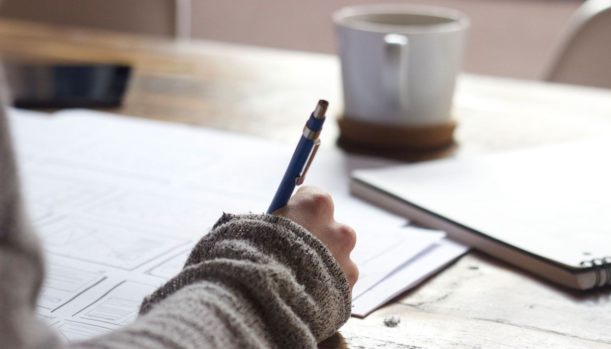 A girl wearing a gray sweater and writing in a paper with a cup of coffee in front