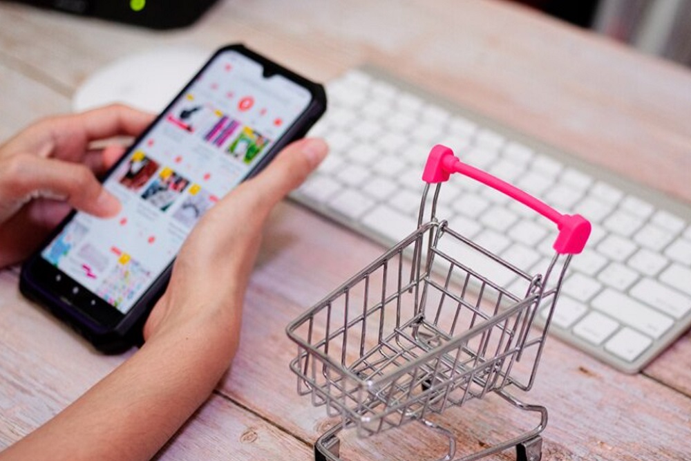 Hands scrolling on a phone in front of a laptop and shopping cart on the side
