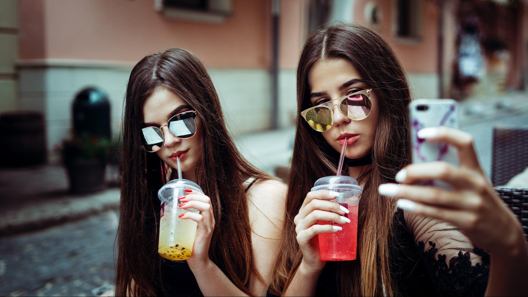 Two woman wearing a sunglasses while taking a selfie and sipping their drinks