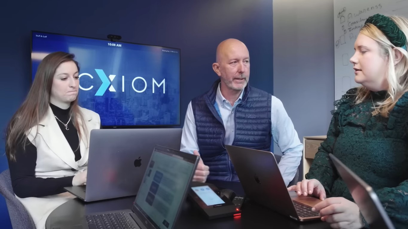 Acxiom CEO Chad Engelgau flanked by two female Acxiom workers, with the Acxiom logo on a TV screen