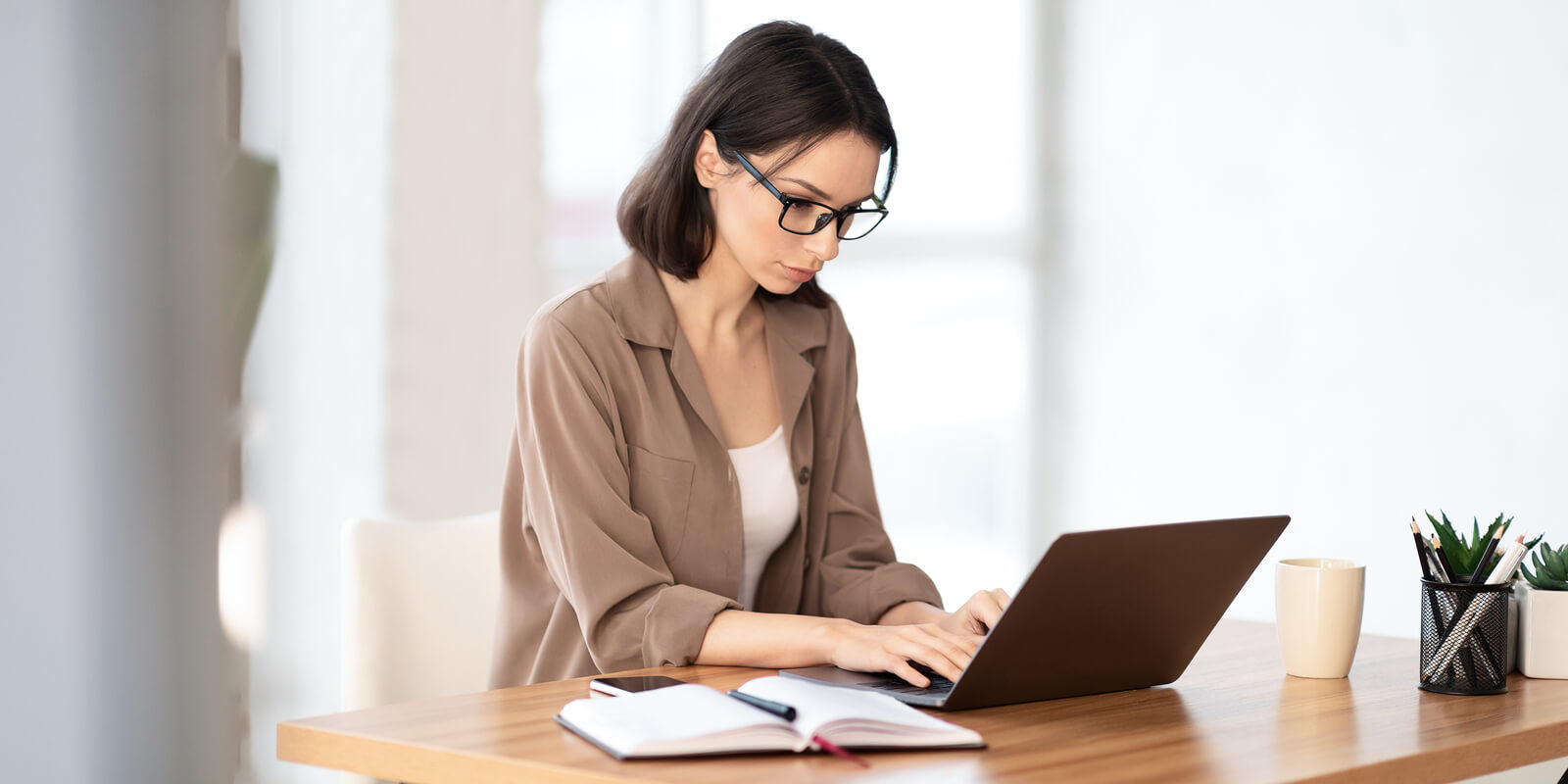 A woman in brown coat and black eyeglasses typing on a laptop