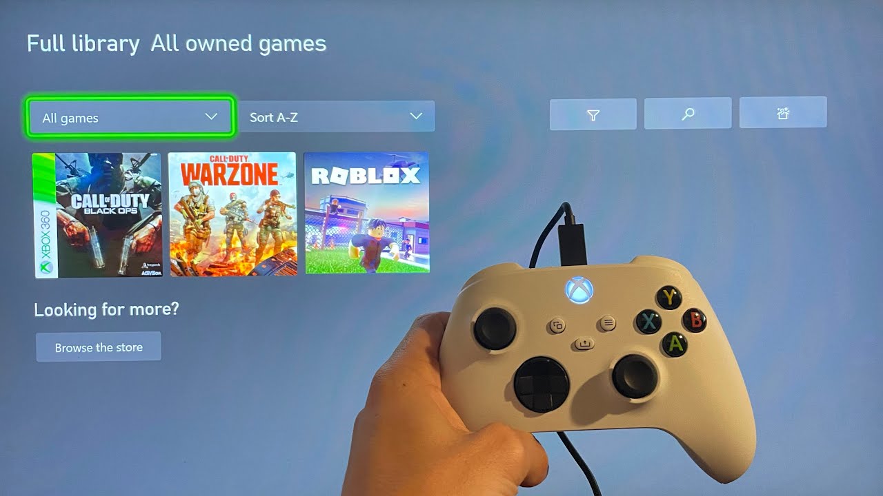 A hand holding a controller on a screen with the XBox game library shown.