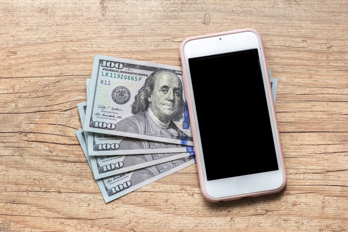 A fan of dollar bills with a phone on top of it on a wooden surface.