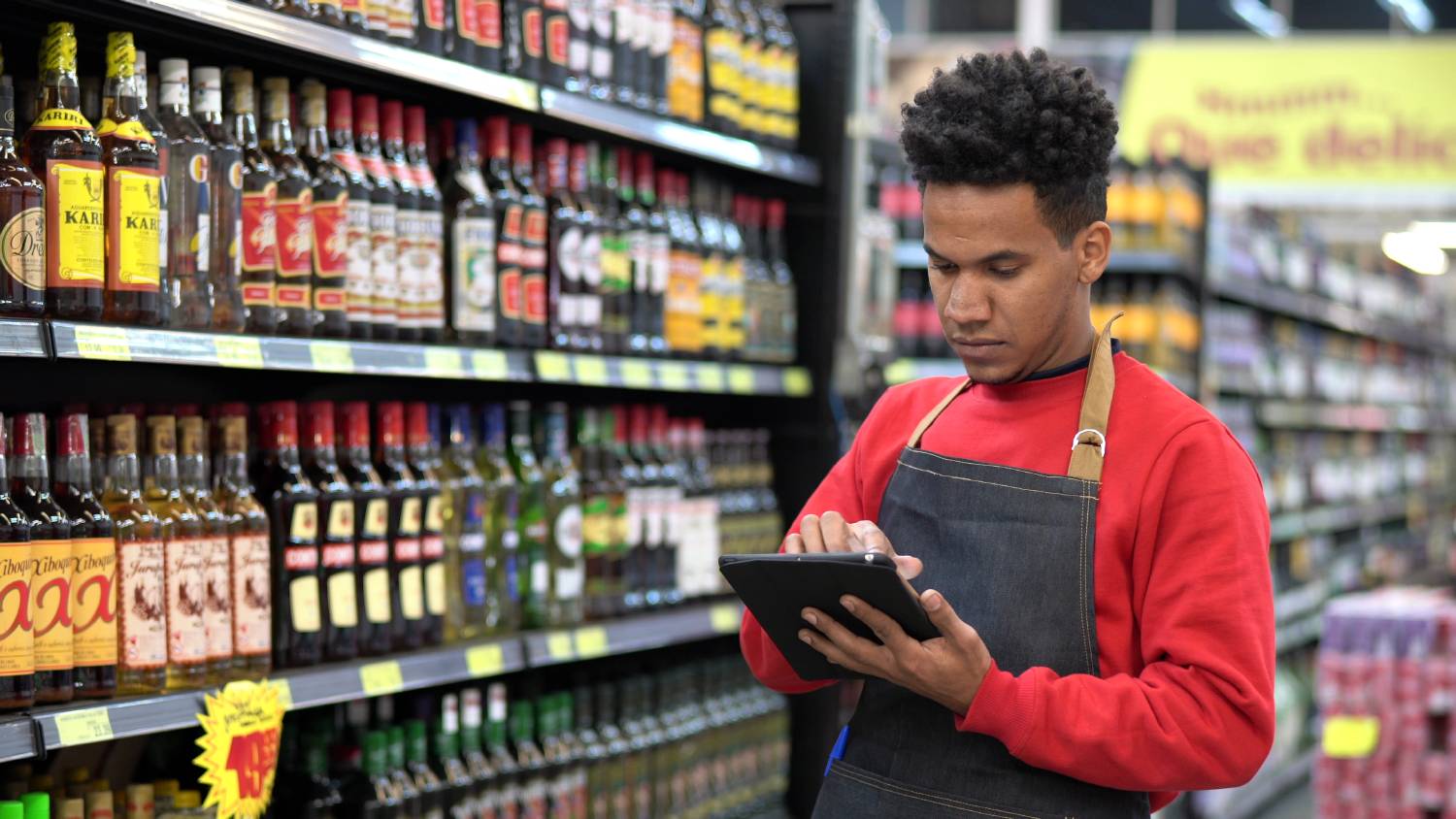 A man wearing a black apron holding a tablet on a grocery aisle