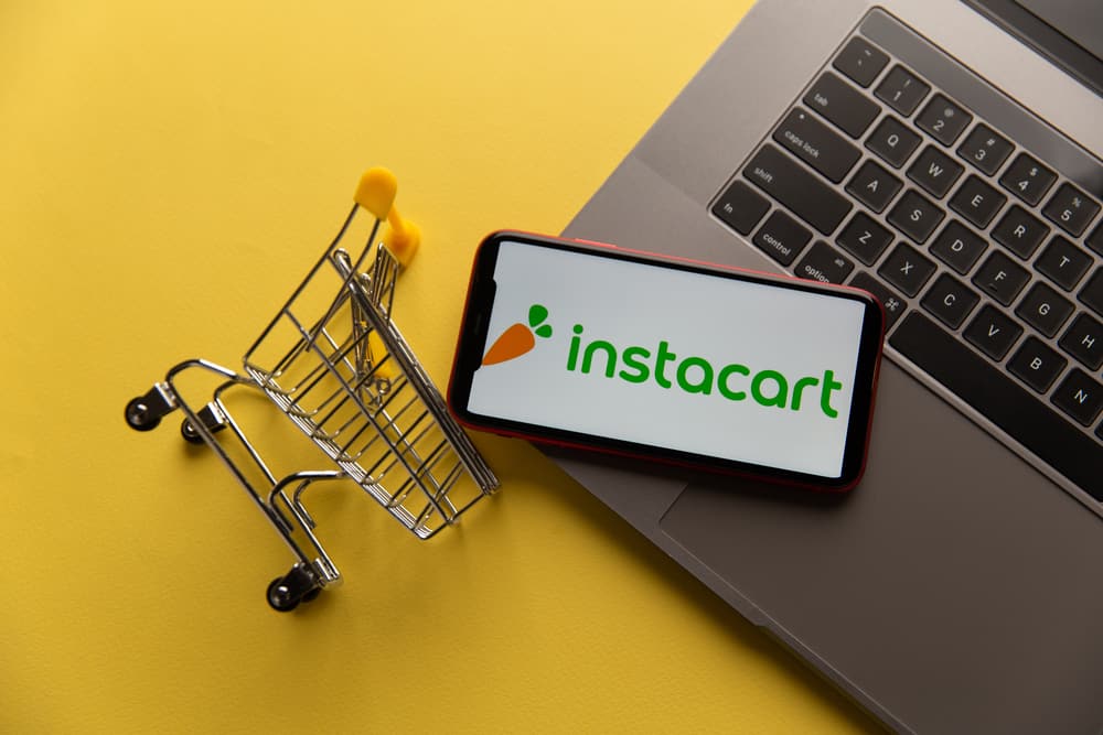 An instacart logo on a phone with small shopping cart on a laptop