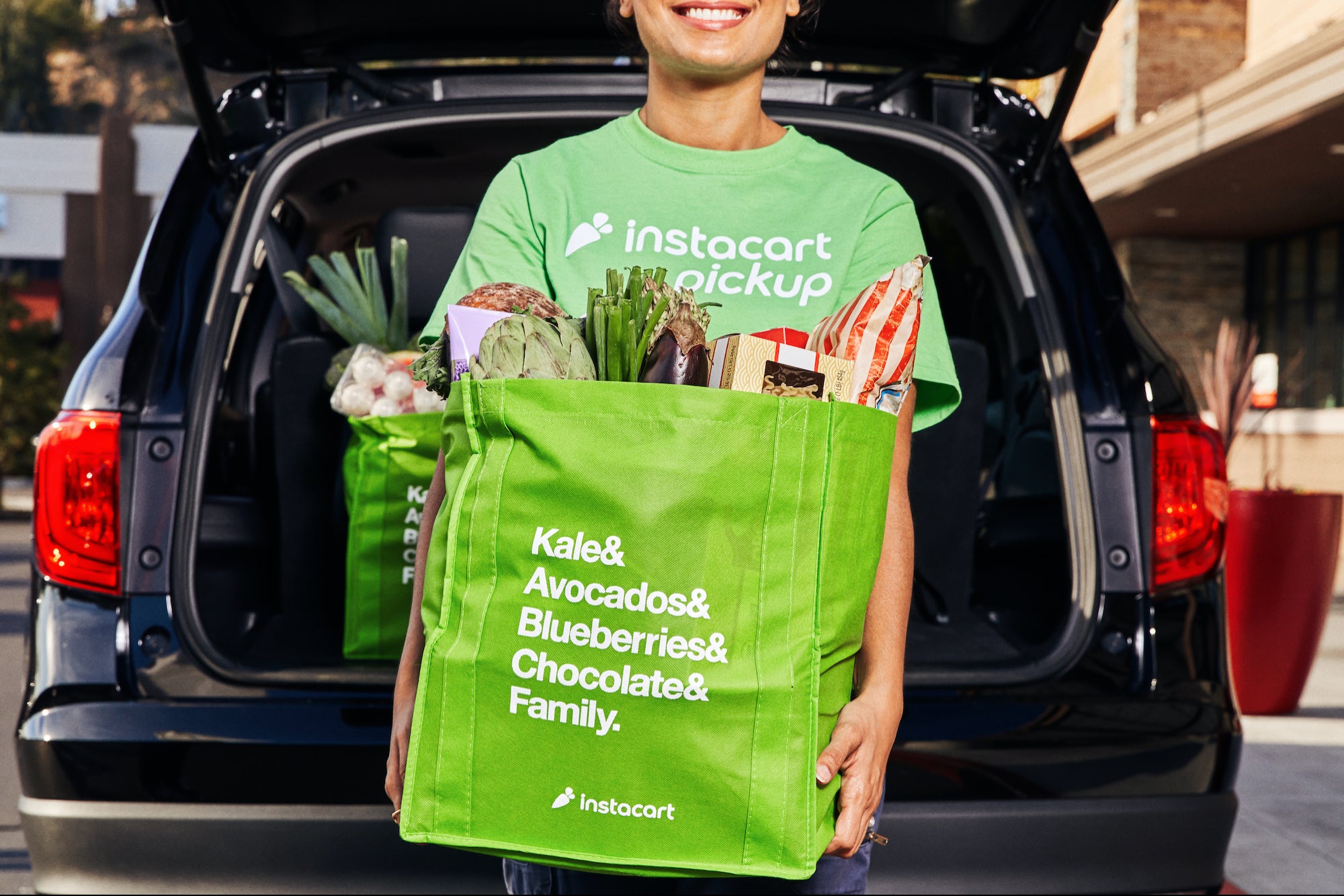 A person holding a green grocery bag with car behind him
